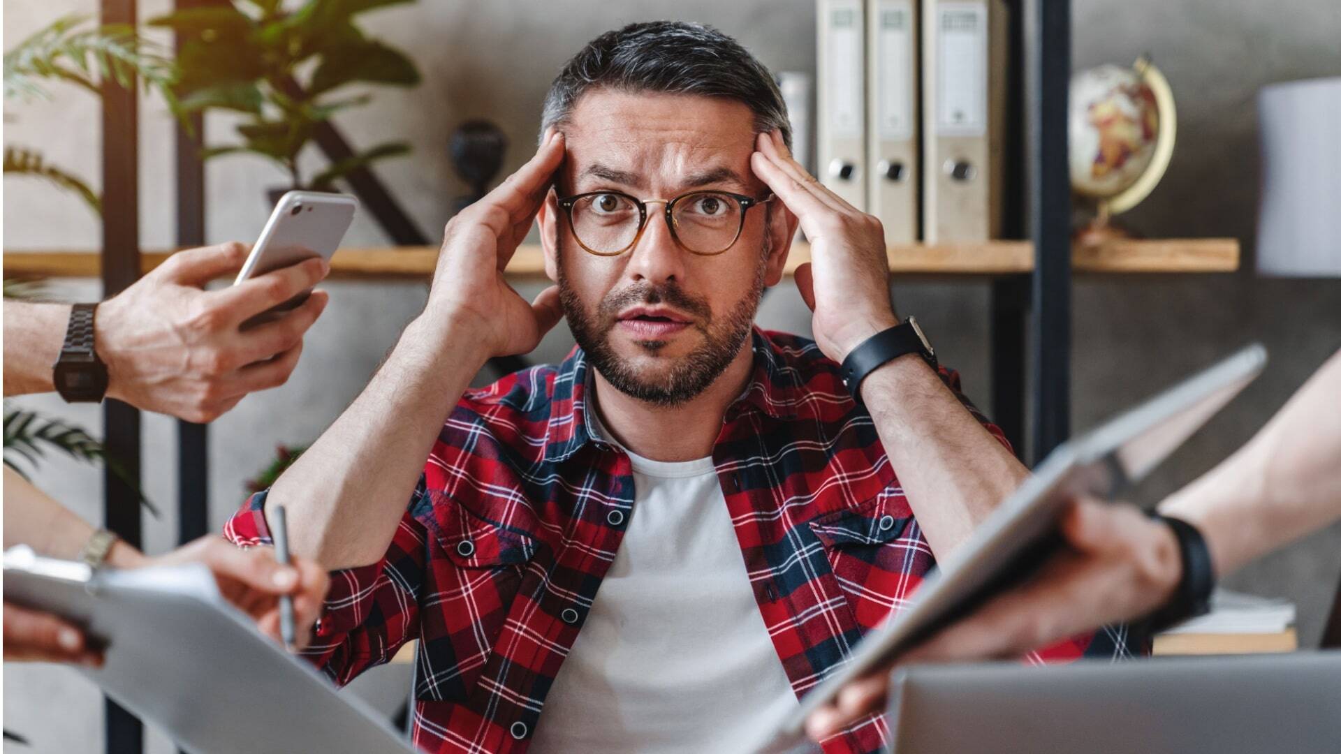 A stressed man in a red plaid shirt sits at a desk, surrounded by hands showing a phone, tablet, and clipboard. This represents the overwhelm of content marketing for a coaching business.